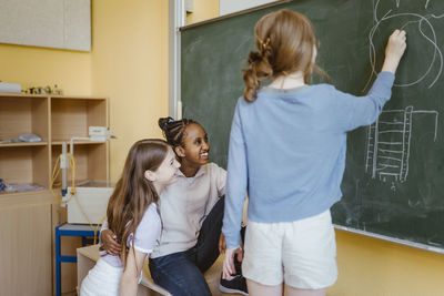 Girls having fun with female friend drawing on chalkboard in classroom