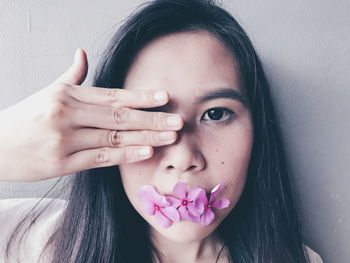 Close-up portrait of woman with pink flowers in mouth covering eye by wall