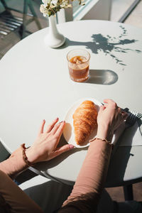 Anonymous female in beret sitting at table in cafe with aromatic glass of coffee and freshly baked croissant