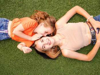 Two girls lie on the grass and laugh, one girl whispers in the ear of the second girl and she smiles