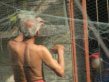 Rear view of shirtless man looking through chainlink fence