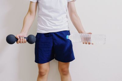 Midsection of man holding dumbbell and water bottle while standing against white background