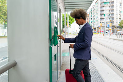 Black man with afro hair pulling out a ticket on the tram with a suitcase next to him and a wireless headset around his neck