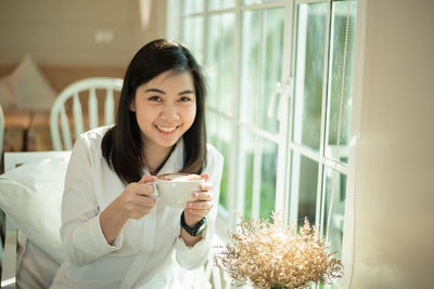 Portrait of young woman drinking coffee while sitting by window at cafe