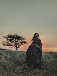 Woman with blanket standing on grassy land against clear sky during sunset