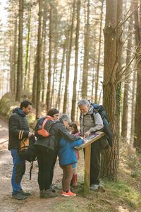 Family of hikers with kids reading information on wooden board while standing on path in woods in autumn during trekking