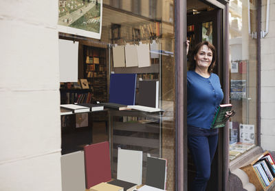 Portrait of smiling librarian holding book while standing at doorway