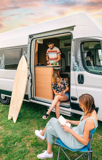 Friends traveling in a camper van. man cooking, woman reading and woman looking mobile