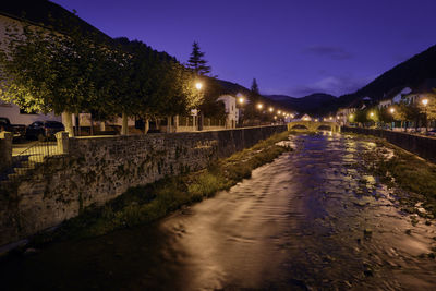 Illuminated street by buildings against sky at night