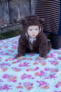 A small child in a brown teddy bear overalls sits on a blanket , adult stays behind him.