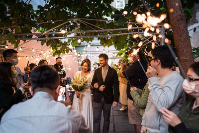 Group of people with lit sparklers congratulating bride and groom during wedding celebration on street decorated with lightbulb garlands