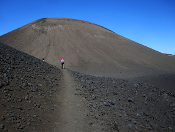 Rear view of man walking on dirt road against clear blue sky