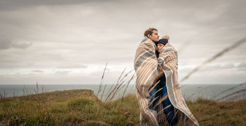 Couple covered with blanket standing by sea on field