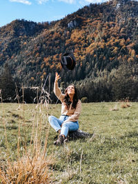 Young woman sitting on grass in valley, throwing hat in air, autumn, fall, happy.