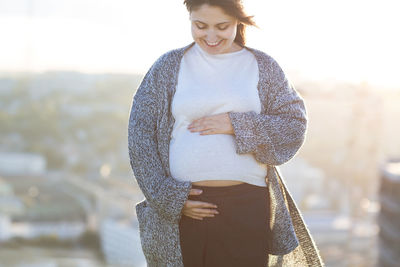 Smiling pregnant woman with hands on stomach standing against clear sky