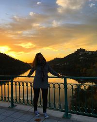 Rear view of woman standing on railing against sky during sunset