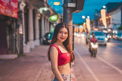 Portrait of smiling young woman standing on street in city at dusk