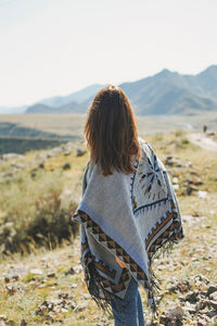 Brunette young woman traveler in poncho from back on road, trip to mountains, altai