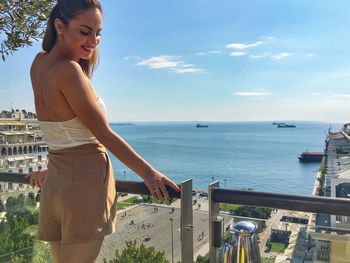 Rear view of young woman standing at balcony with sea in background against blue sky