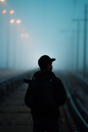 REAR VIEW OF MAN STANDING BY RAILROAD TRACKS AGAINST SKY
