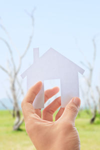 Cropped hand of person holding paper house against sky