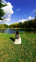 Rear view of woman sitting on grass by lake against sky