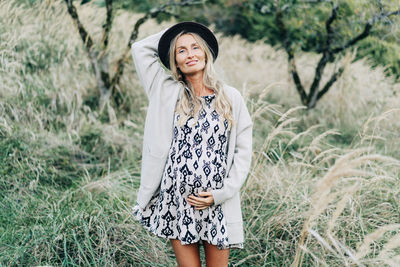 Beautiful happy pregnant woman standing in the park wearing a dress and hat.