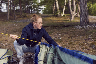 Young woman crouching while setting up tent against trees at campsite