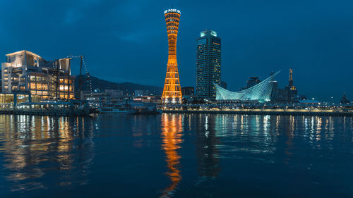 Illuminated modern buildings by river against sky at dusk