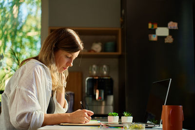 Woman working at home, writing in notebook and using laptop