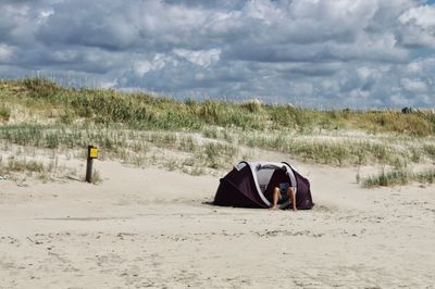 Man lying down in tent on sand dunes at beach against sky