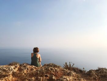 Woman sitting on rock looking at sea against clear sky
