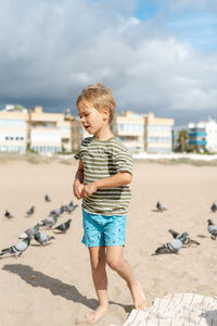 A boy feeds pigeons on a sandy beach against a background of apartment complexes and a stormy sky