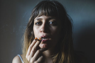 Close-up portrait of young woman smoking cigarette against gray background