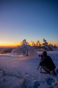 Man taking a photo on snow covered landscape against clear sky during sunset