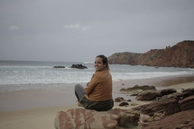 Portrait of young woman sitting on rock at beach against sky