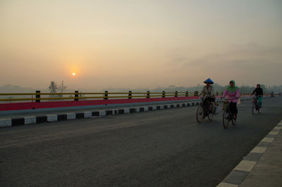 People riding bicycle on road against sky during sunset