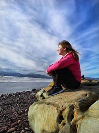 Girl sitting on rock at beach against sky