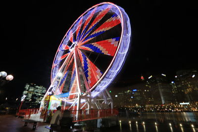 Illuminated ferris wheel by buildings against sky at night