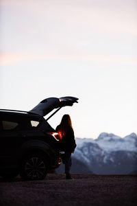 Back view of anonymous traveling female standing near modern automobile and admiring peaceful scenery of mountains in evening