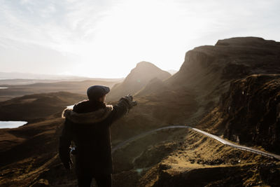 Back view of unrecognizable male tourist in outerwear standing on hill and observing spectacular view of mountains in autumn in scottish highlands while pointing away