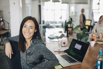 Portrait of smiling female computer programmer sitting at desk in creative office