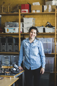 Portrait of confident mature female teacher standing by science project at desk in classroom at high school