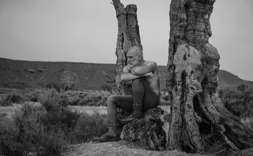 Monochrome of shirtless man sitting on rock by bare olive tree in desert against sky