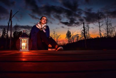 Young woman sitting by tree against sky at sunset