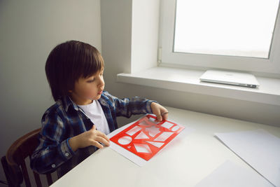 Boy child draws on paper with a ruler on a table sitting by the window