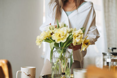 Young woman in white shirt with bouquet of yellow flowers in hands near window at home
