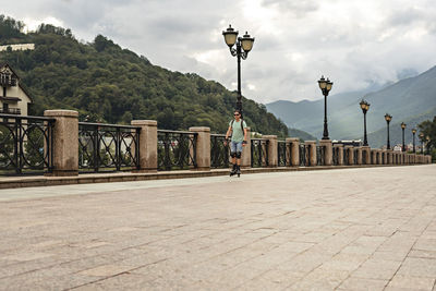 Young man riding on roller skates along embankment against background of forest and mountains 