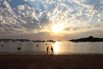 Boys playing on shore at beach against cloudy sky during sunset