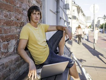 Young man with laptop sitting on sit against brick wall